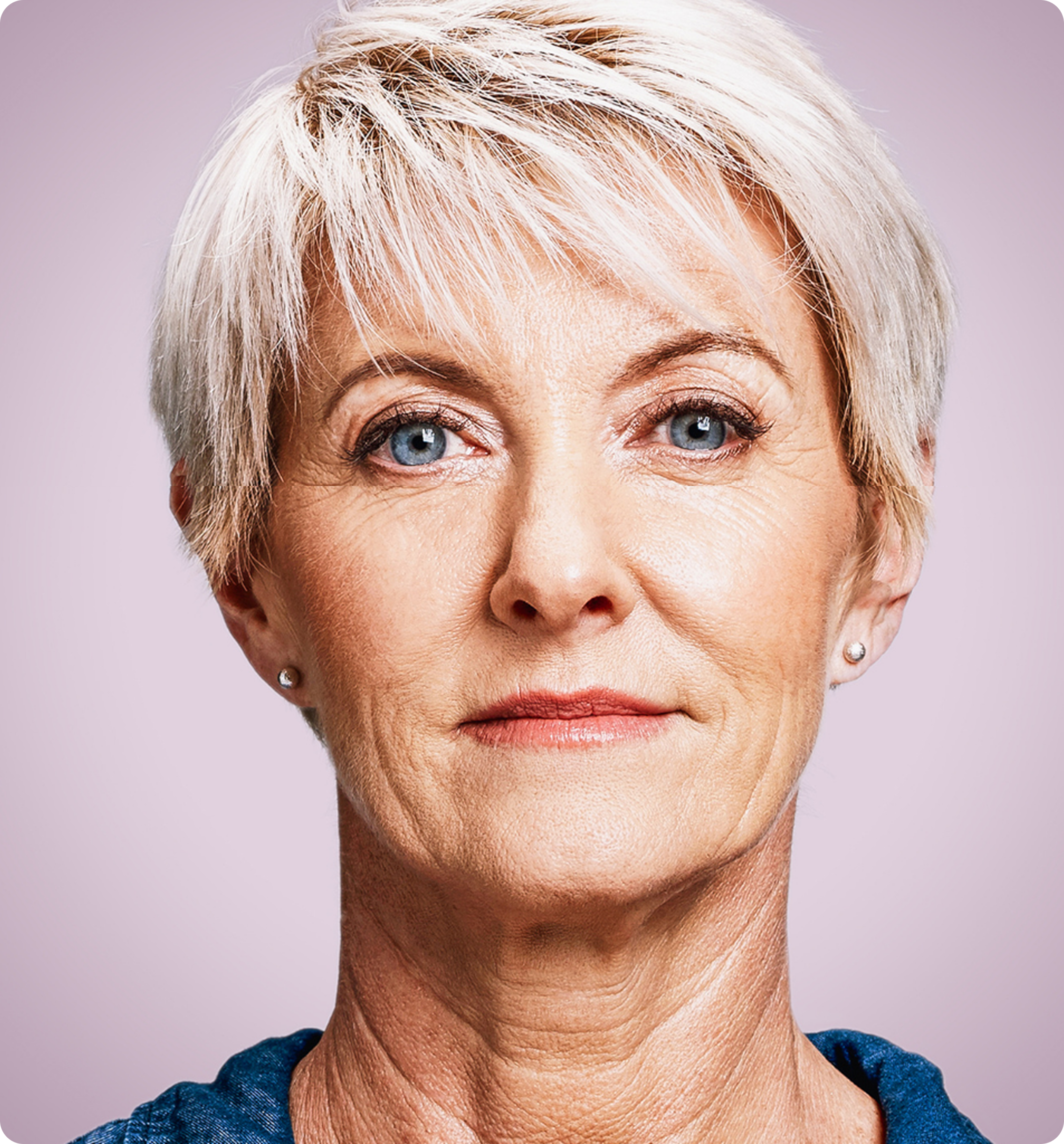 Eine Frau mit kurzen, weißen Haaren und blauen Augen blickt vor einem schlichten, hellvioletten Hintergrund direkt in die Kamera. Sie trägt ein blaues Oberteil und kleine Ohrstecker.