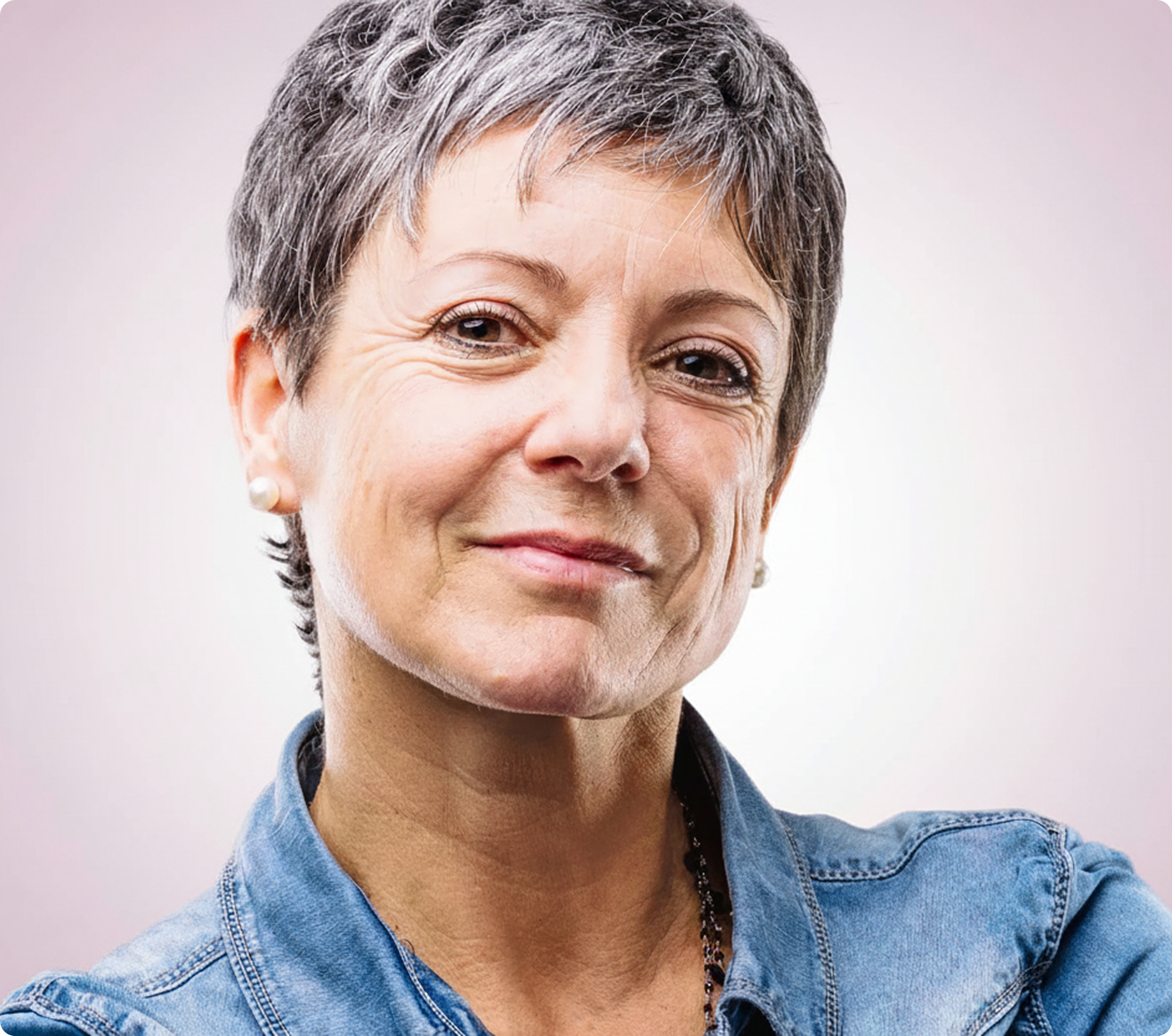 Eine Frau mit kurzen grauen Haaren und einem Jeanshemd blickt mit einem leichten Lächeln in die Kamera, vor einem schlichten hellen Hintergrund.