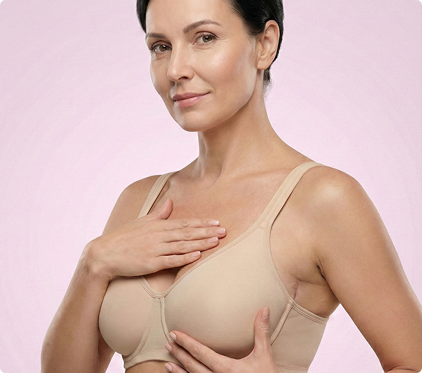 Eine Frau, die einen beigen BH trägt, legt eine Hand auf ihre Brust und die andere unter ihre Brust, während sie vor einem hellrosa Hintergrund steht.