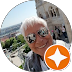 Eine Person mit kurzen weißen Haaren und Sonnenbrille macht ein Selfie im Freien, mit historischer Architektur und einer Stadtansicht im Hintergrund. In der Ecke erscheint ein orangefarbener Stern.