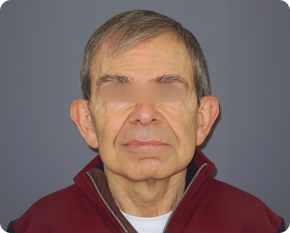 Älterer Mann mit kurzen grauen Haaren, der einen kastanienbraunen Pullover mit Reißverschluss über einem weißen Hemd trägt und vor einem schlichten grauen Hintergrund nach vorne schaut. Die Augen sind zum Schutz der Privatsphäre unscharf.