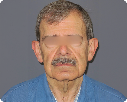 Ein älterer Mann mit grauem Haar und Schnurrbart, der ein blaues Hemd mit Kragen trägt, blickt nach vorne auf einen schlichten grauen Hintergrund. Seine Augen sind aus Gründen der Privatsphäre unscharf.