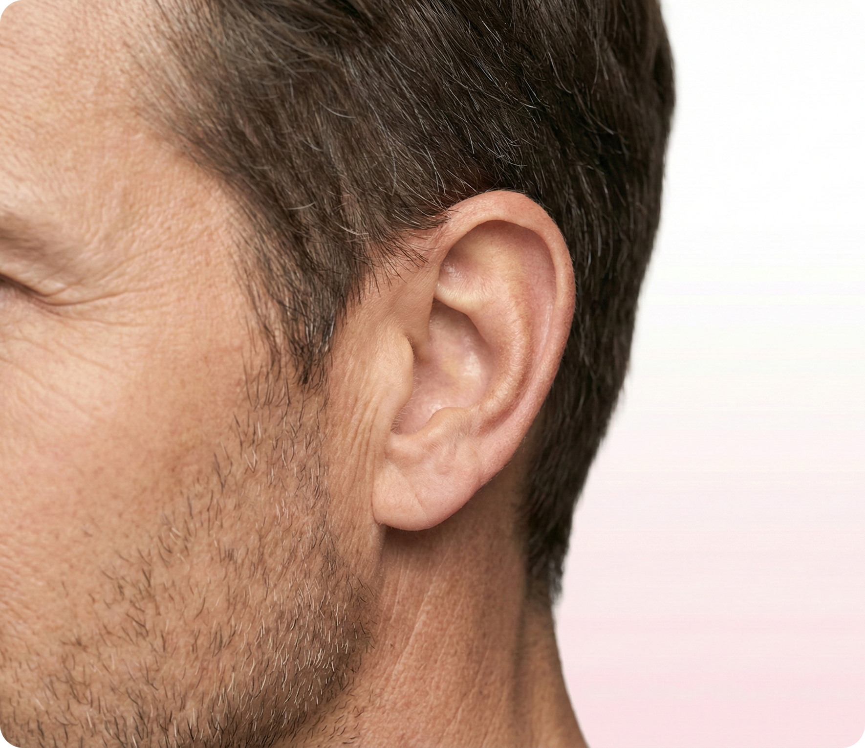 Nahaufnahme der linken Gesichtshälfte eines Mannes mit kurzen braunen Haaren und leichten Bartstoppeln vor einem blassen Hintergrund, wobei das Ohr im Mittelpunkt steht.