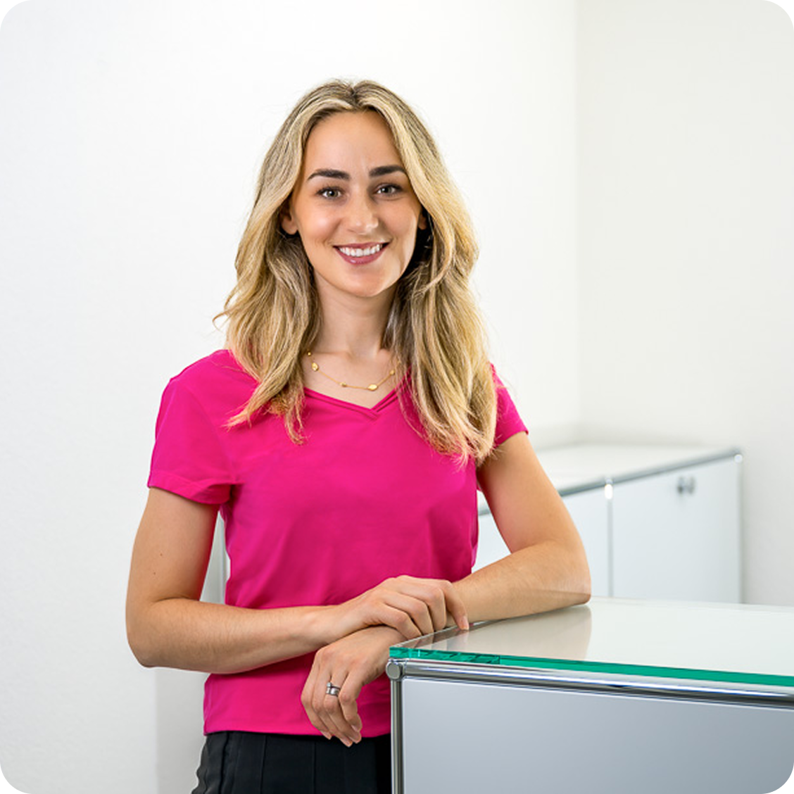 Eine Frau mit langen blonden Haaren und einem leuchtend rosafarbenen Oberteil steht lächelnd an einem gläsernen Tresen in einem modernen, weißen Büroraum.