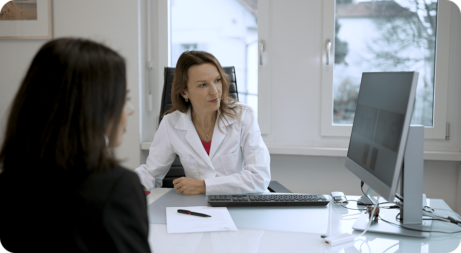 Eine Ärztin in einem weißen Kittel sitzt an einem Schreibtisch und schaut auf einen Computerbildschirm, während sie mit einem Patienten gegenüber spricht. Auf dem Schreibtisch befinden sich eine Tastatur, eine Maus und Papierkram.