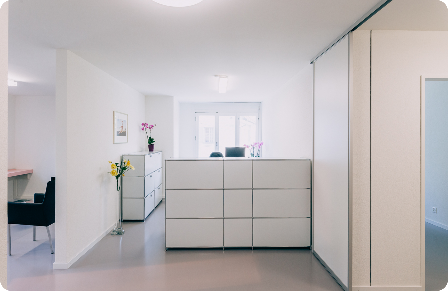 Helle, moderne Büroräume mit weißen Wänden, Aktenschränken, Blumen in Vasen, einem Schreibtisch mit zwei Stühlen und großen Fenstern, die natürliches Licht hereinlassen.