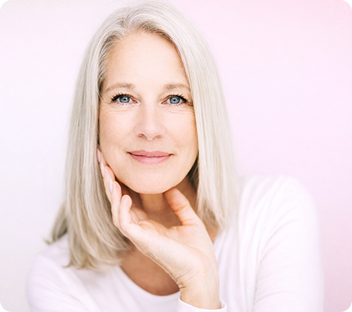 Eine Frau mit langen grauen Haaren und blauen Augen, die ein weißes Oberteil trägt, stützt ihr Kinn auf ihre Hand und lächelt sanft vor einem hellrosa Hintergrund.