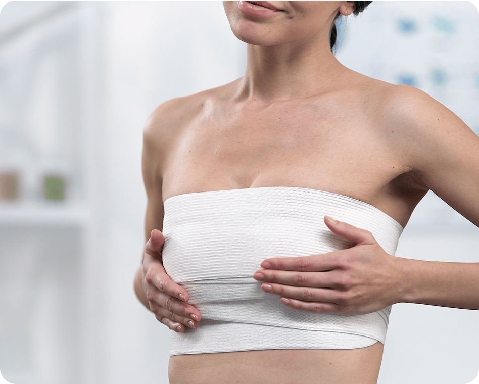 Eine Frau mit einem weißen Bandeau-Top hält sich mit beiden Händen die Brust, während sie in einem schwach beleuchteten Innenraum steht.