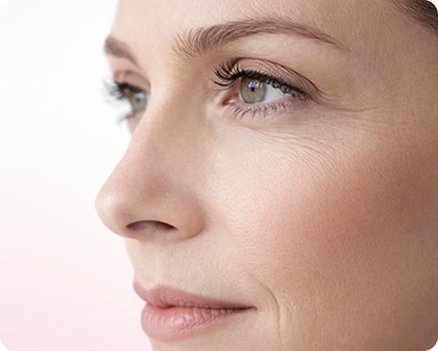 Nahaufnahme des Gesichts einer Frau im Profil mit klarer Haut, grünen Augen, leichtem Make-up und neutralem Gesichtsausdruck vor einem weichen, hellen Hintergrund.
