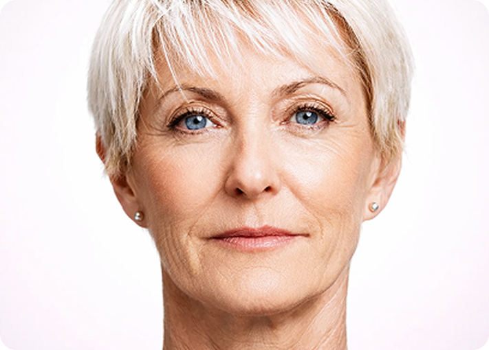 Nahaufnahme einer älteren Frau mit kurzen, weißen Haaren, blauen Augen und heller Haut, die mit neutralem Blick direkt in die Kamera schaut.