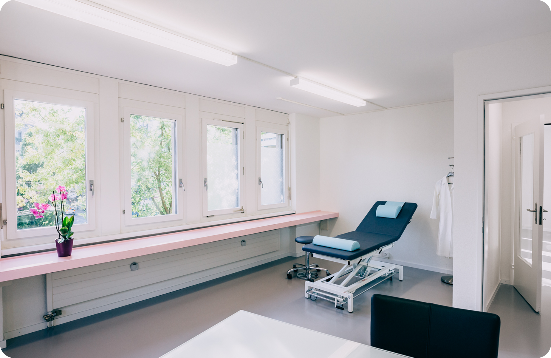 Ein sauberer, heller medizinischer Untersuchungsraum mit einem Untersuchungstisch, Fenstern, einer Topfpflanze und einem weißen Kittel an der Wand.