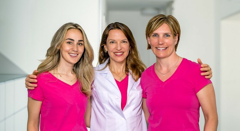 Drei Frauen stehen in einem Innenraum zusammen; zwei von ihnen tragen passende rosa Hemden, und die Frau in der Mitte trägt einen weißen Laborkittel über einem rosa Oberteil. Sie lächeln in die Kamera.