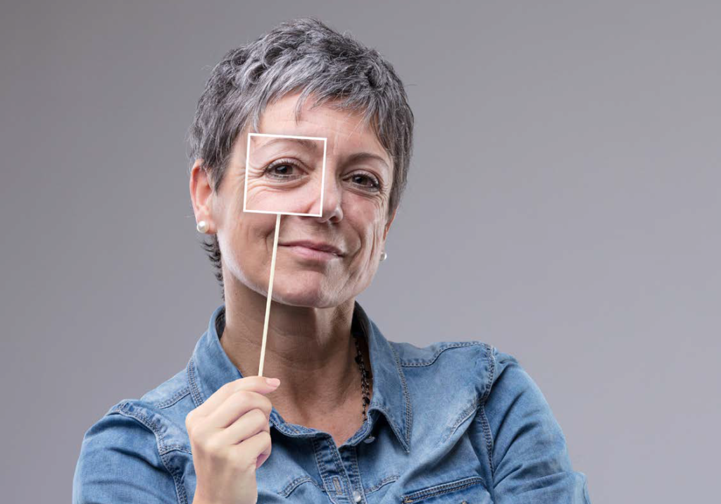 A woman with short grey hair holds a square frame on a stick over her eye. She is standing in front of a plain grey background and is wearing a denim shirt.