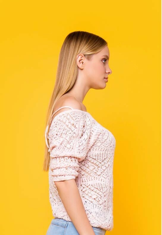 Frau mit langem glattem blondem Haar und weißem Strickpullover steht im Profil vor einem gelben Hintergrund.