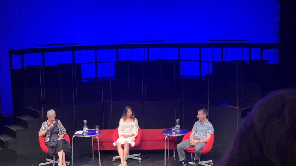 Three people sit on stage in red chairs with small tables and water pitchers, speaking or preparing for a speech in front of a blue-lit background and black scenery.